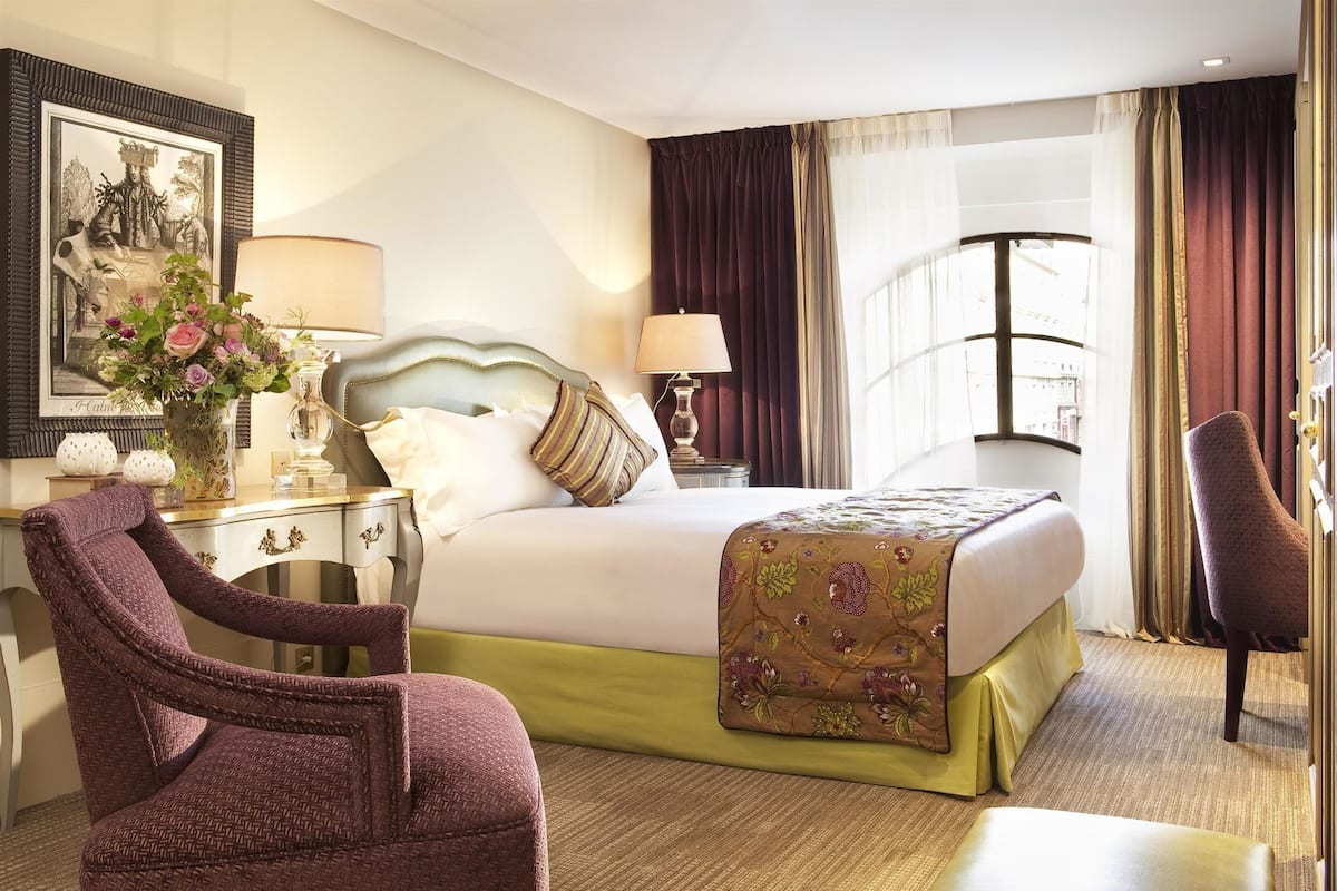 A hotel room with a bed, two bedside tables, a chair, a desk, and a window with curtains.