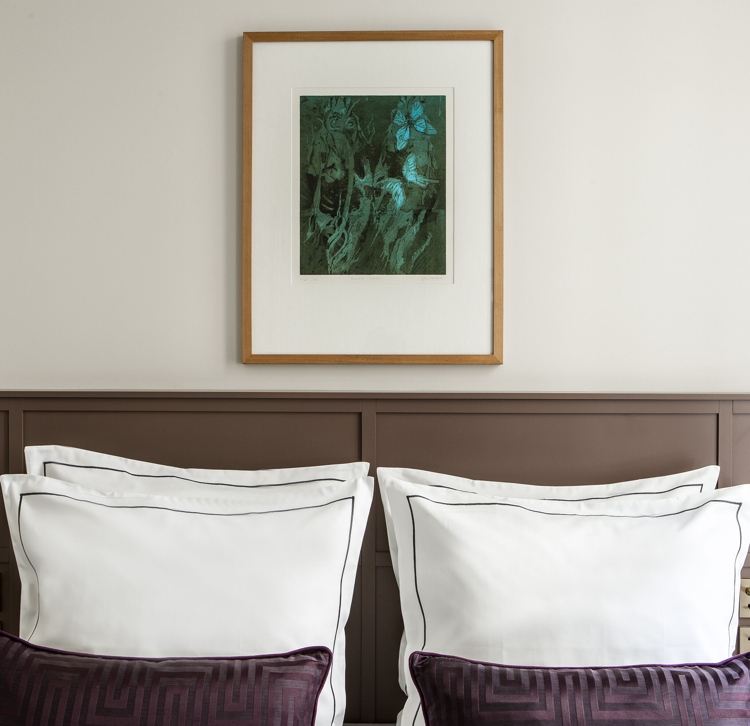 Une œuvre d’art encadrée est accrochée au-dessus d’un lit agrémenté d’oreillers blancs et violets.