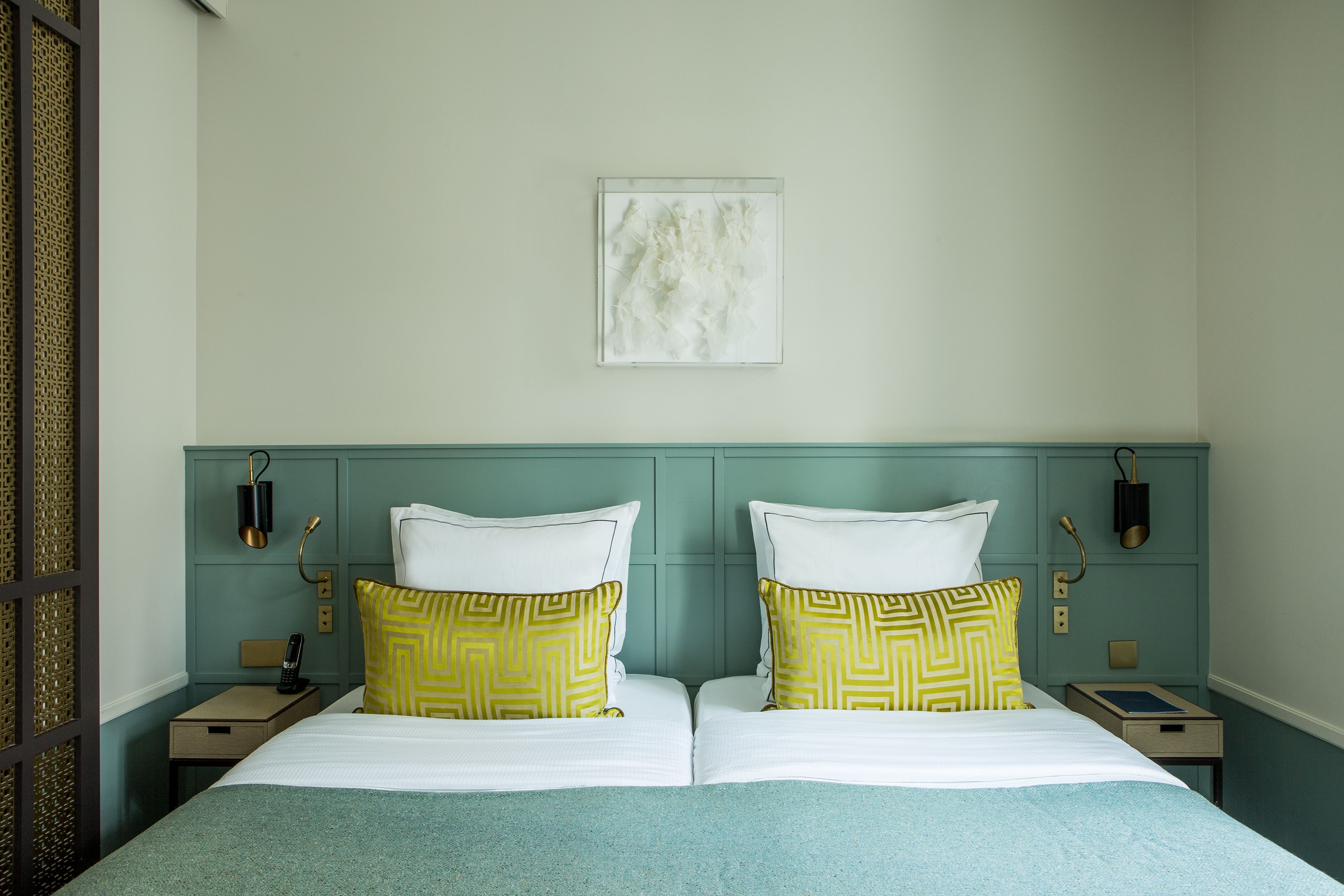 Un lit double avec des oreillers à motifs blancs et jaunes, une tête de lit de couleur turquoise et une petite table de chevet avec une lampe et un livre.