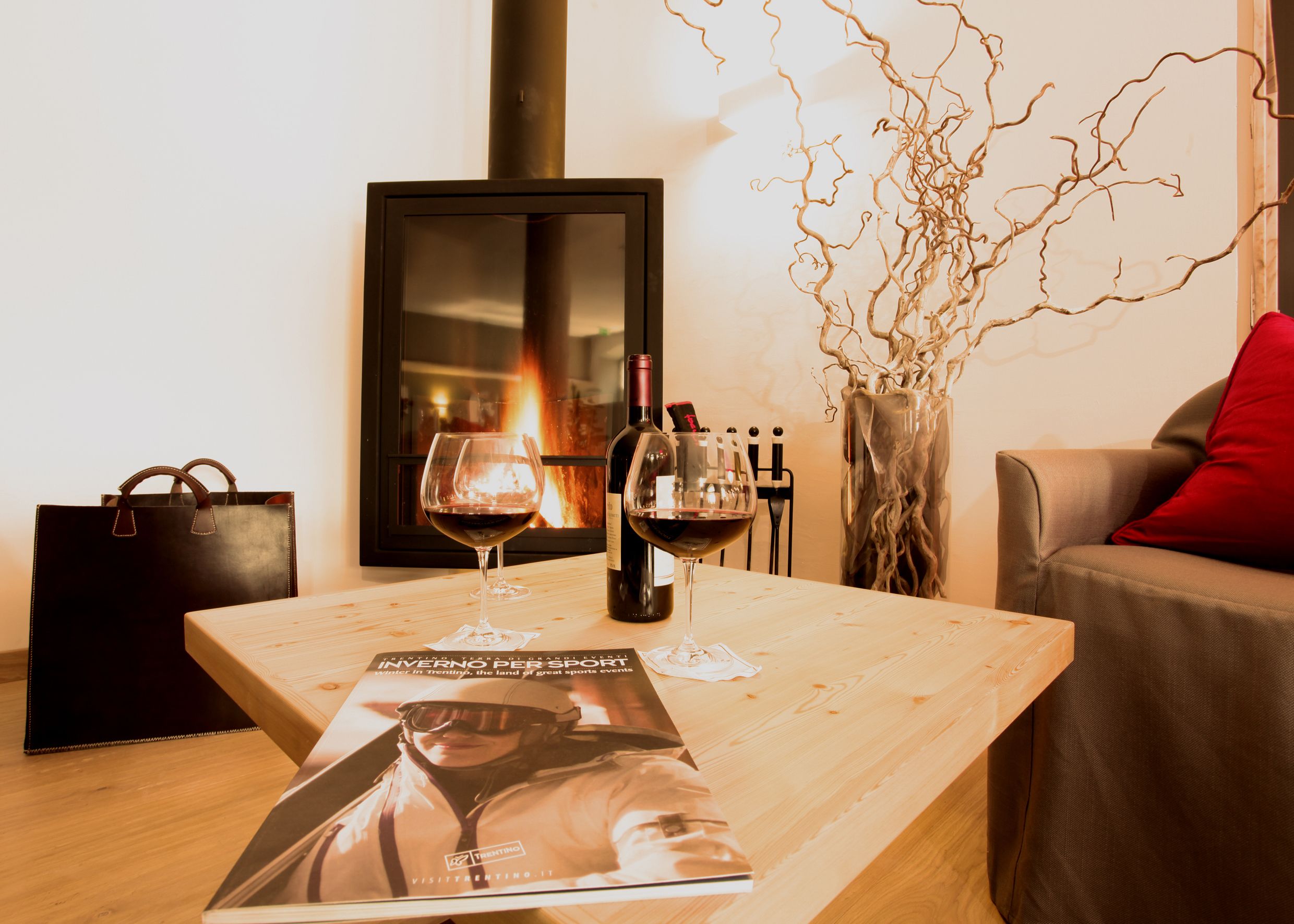 Una mesa de madera con dos copas de vino y una botella, un cuadro enmarcado, un florero con ramas, un bolso negro y una almohada roja sobre un sofá.
