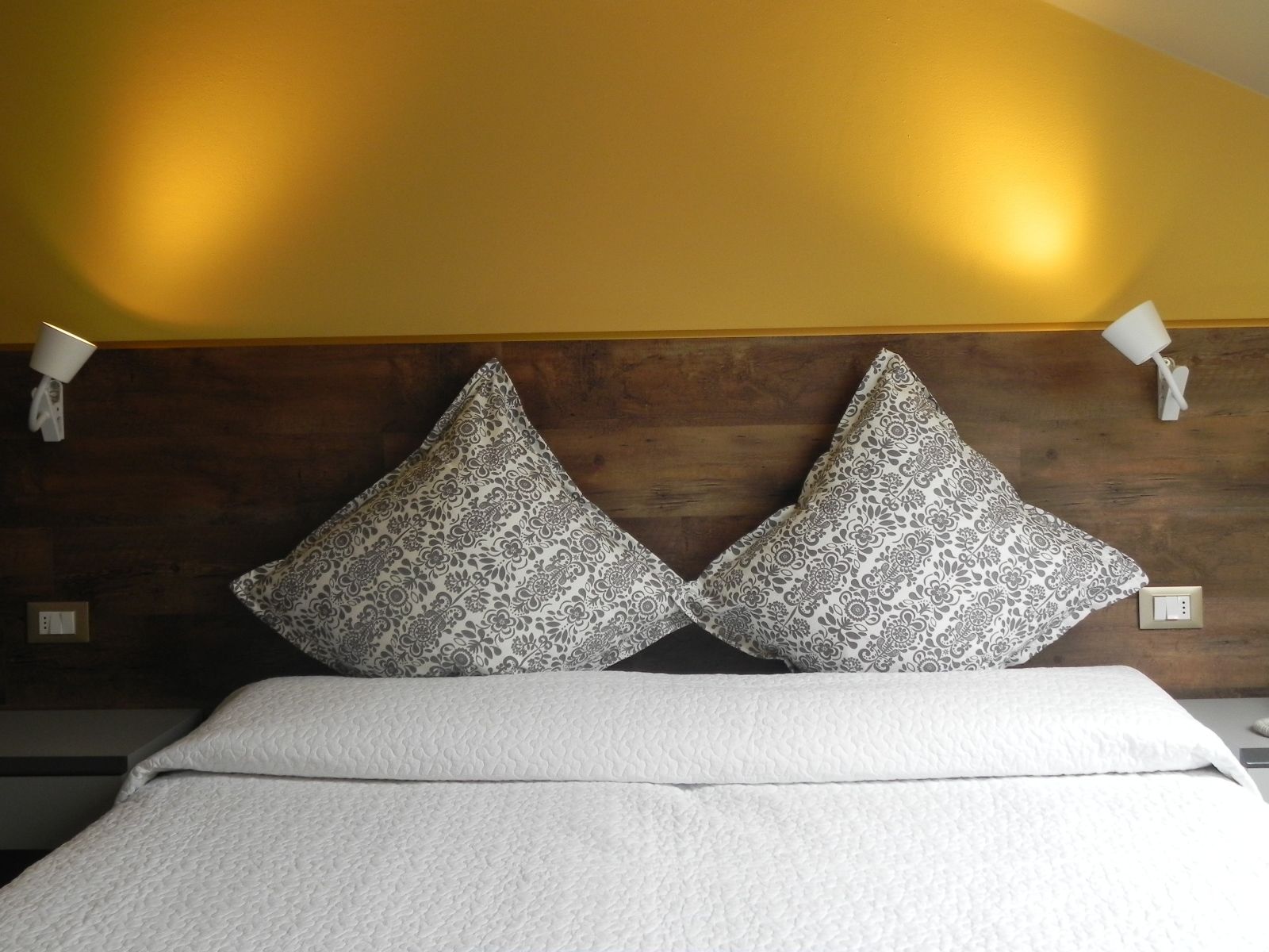 A hotel room with a wooden headboard, two patterned pillows, a white bedspread, and two wall-mounted lamps.
