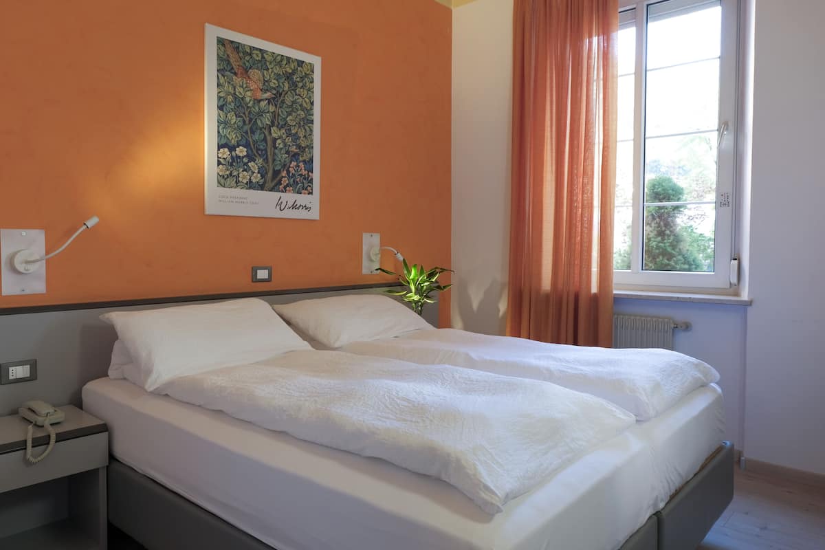 A neatly made bed in a room with a large window, a wall-mounted painting, and a telephone on the nightstand.