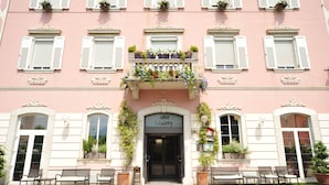 A pink building with a balcony adorned with plants and flowers, a sign reading "hotel Liberty," and benches in front. - Liberty (Levico Terme)