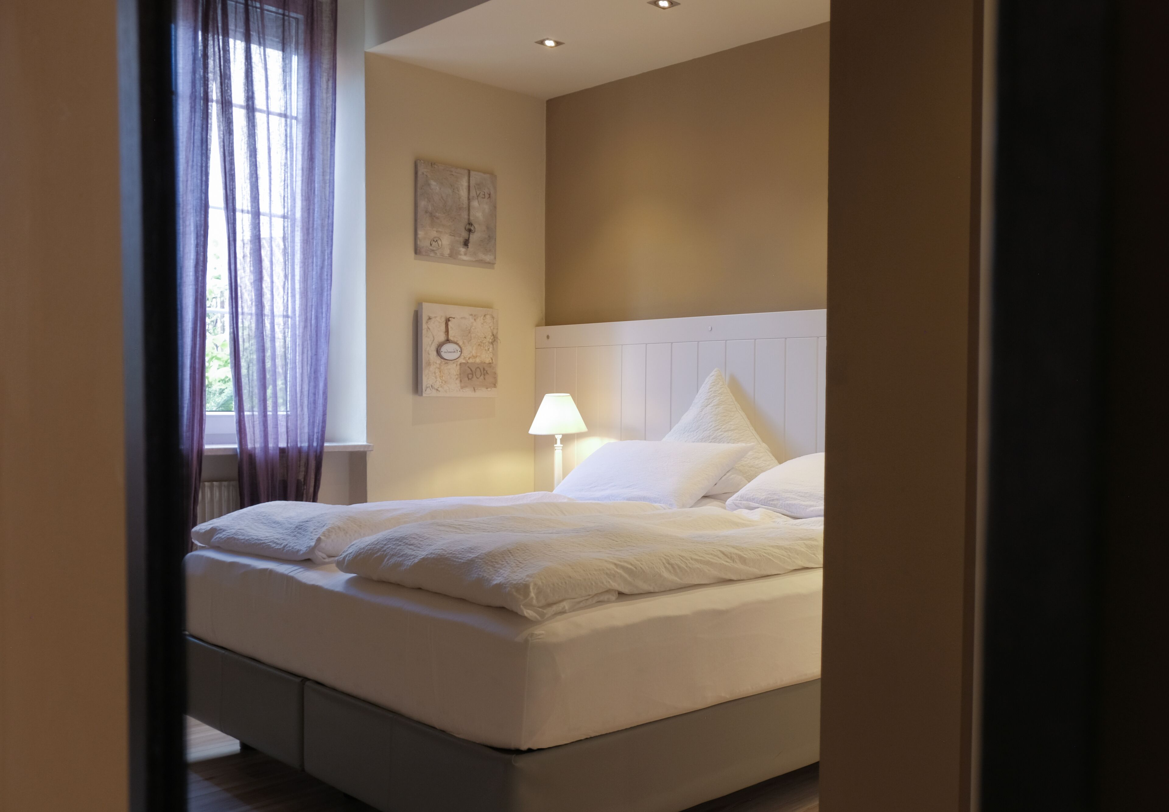Una cama bien hecha con sábanas blancas en una habitación con paredes color beige, una ventana con cortinas traslúcidas y una lámpara de noche.