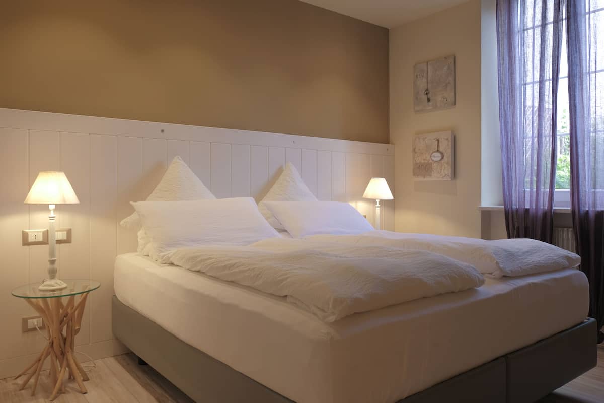 A neatly made bed with white linens in a room with a wooden headboard, two bedside lamps, and a window with sheer curtains.