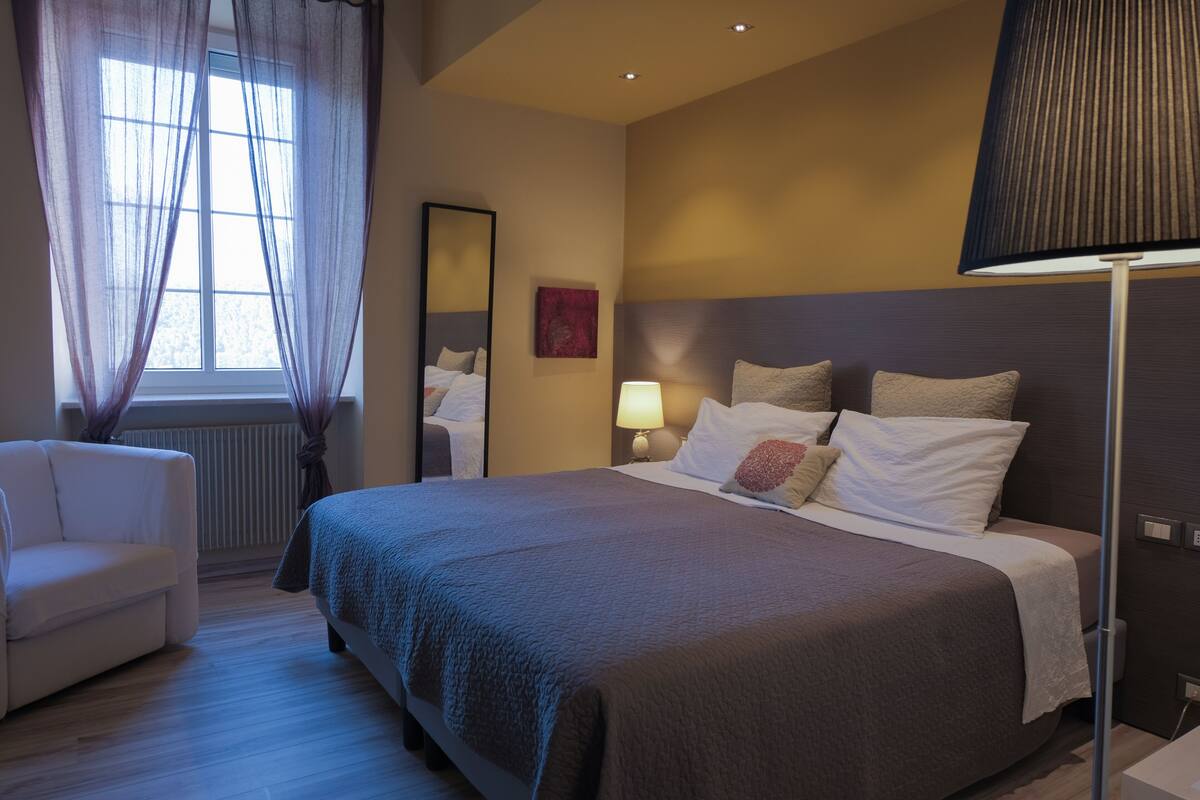 A neatly arranged bedroom with a large bed, a bedside table with a lamp, a chair, and a window with sheer curtains.