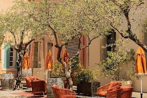An outdoor seating area with wicker chairs and orange umbrellas, surrounded by trees and buildings. - Brit Privilège Victoria Boutique Hotel (Saint-Paul-Trois-Chateaux)