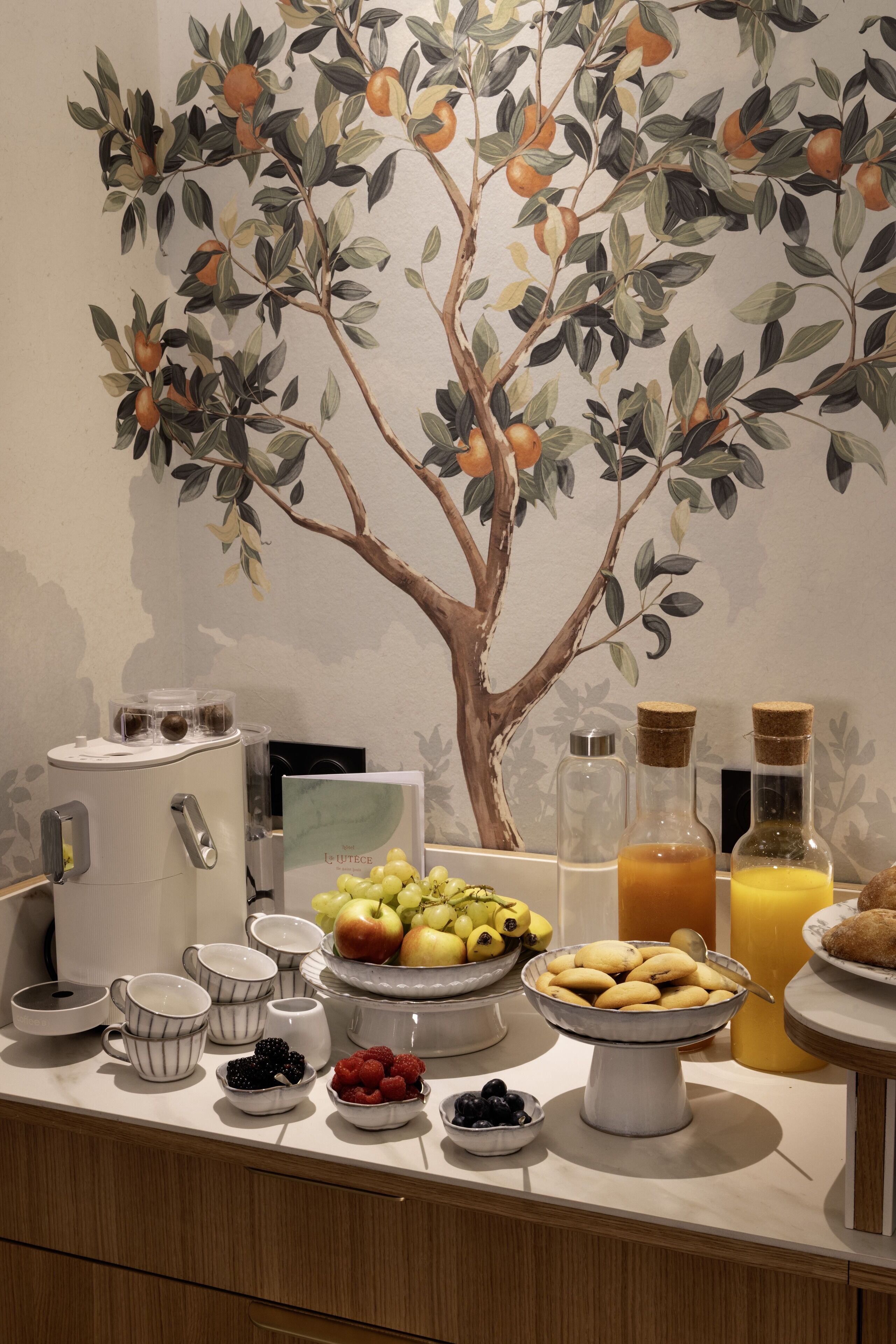 A breakfast buffet with various fruits, pastries, and beverages on a counter with a decorative tree mural in the background.