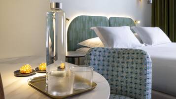 A hotel room with a bed, a chair, a tray with glasses and a bottle, and a wall-mounted lamp.