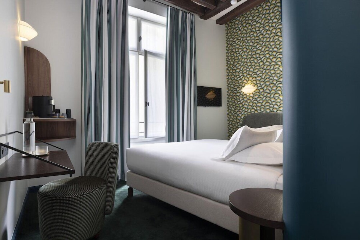 Une chambre d’hôtel comprenant un lit, un bureau, une chaise, une petite table, une machine à café et une fenêtre avec des rideaux.