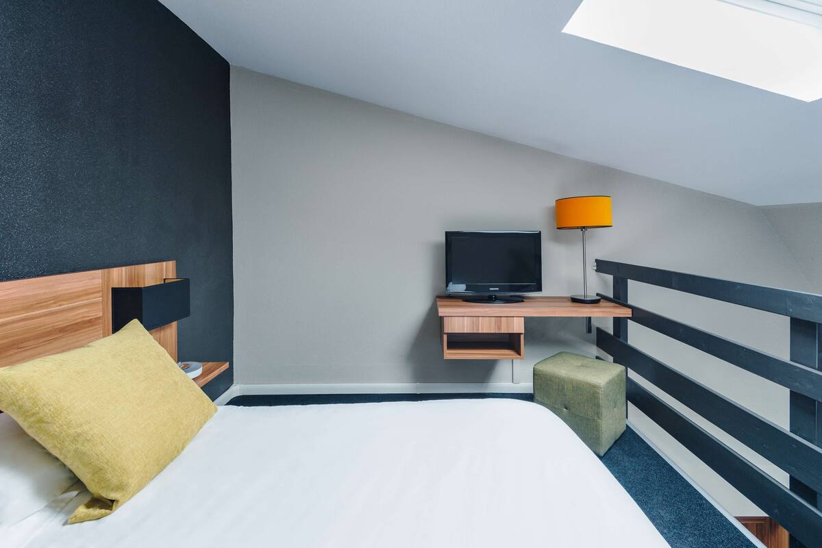 A hotel room with a bed, a TV on a wooden shelf, a yellow pillow, and a green ottoman.