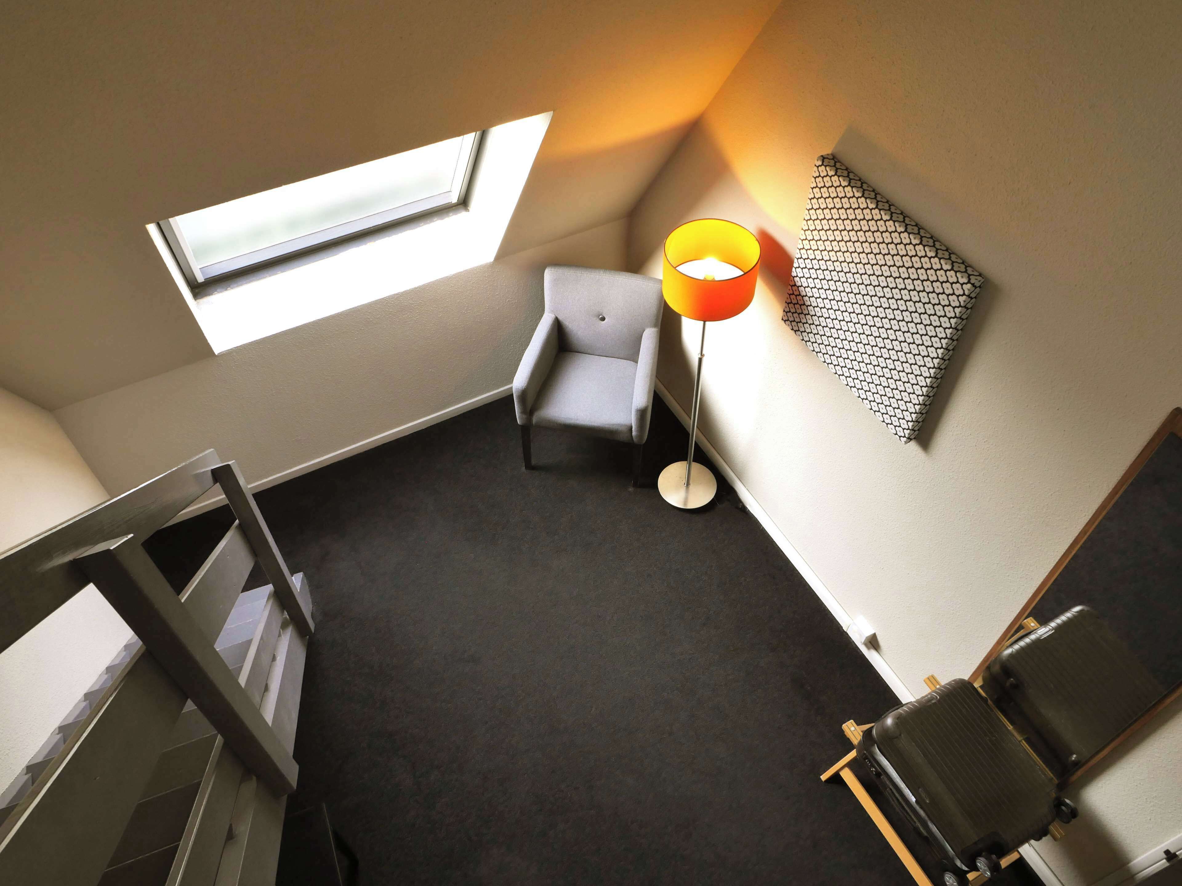 A room with a skylight, a grey chair, a floor lamp, a patterned wall, and a luggage rack.