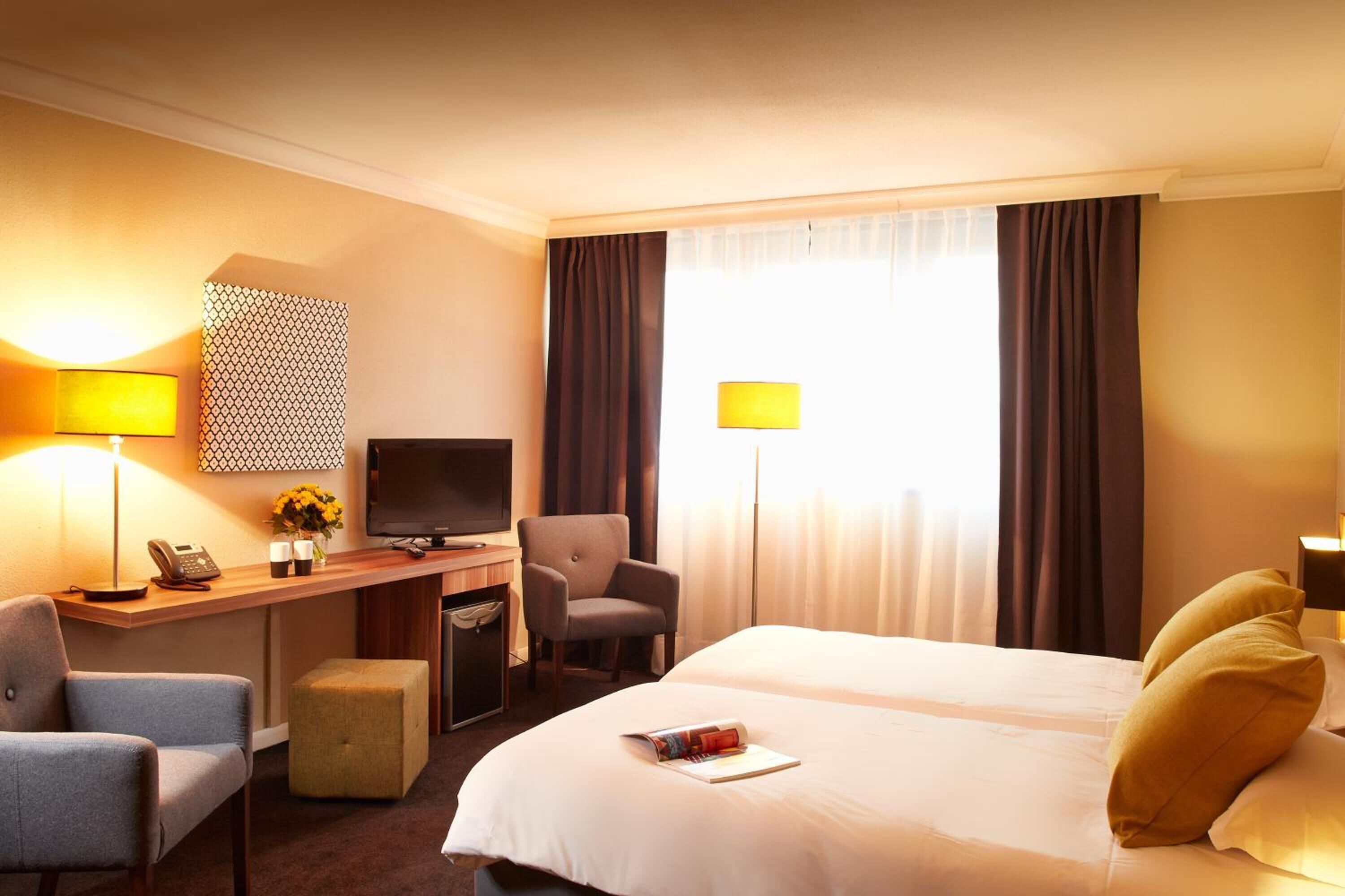 Une chambre d’hôtel comprenant un lit, deux fauteuils, un bureau avec un ordinateur, une télévision et une lampe.