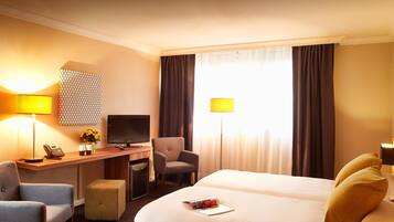 Une chambre d’hôtel comprenant un lit, deux fauteuils, un bureau avec un ordinateur, une télévision et une lampe.