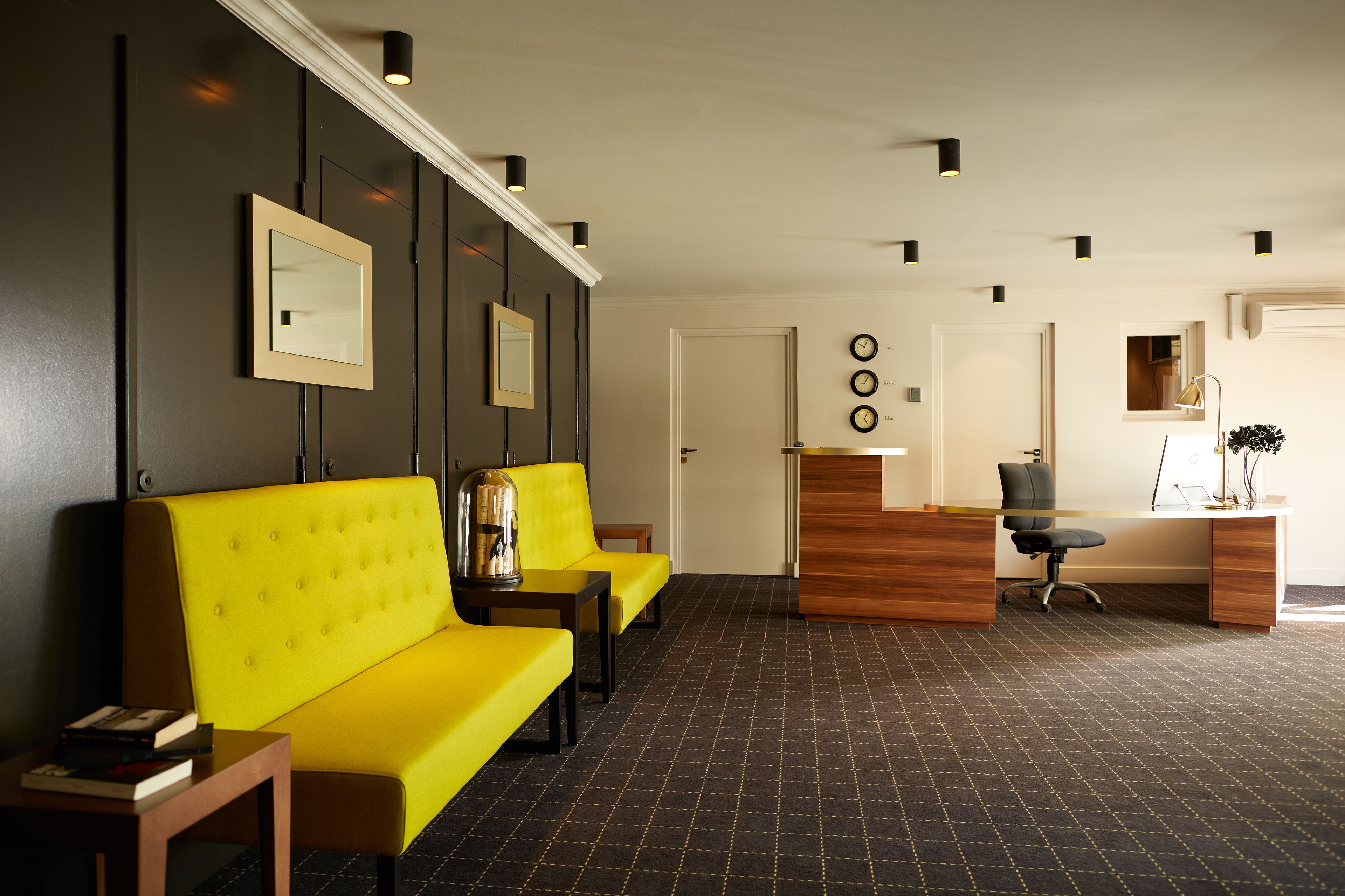 A reception area with a wooden desk, black office chair, and a yellow sofa. There are framed pictures on the wall and a clock.