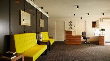 A reception area with a wooden desk, black office chair, and a yellow sofa. There are framed pictures on the wall and a clock.