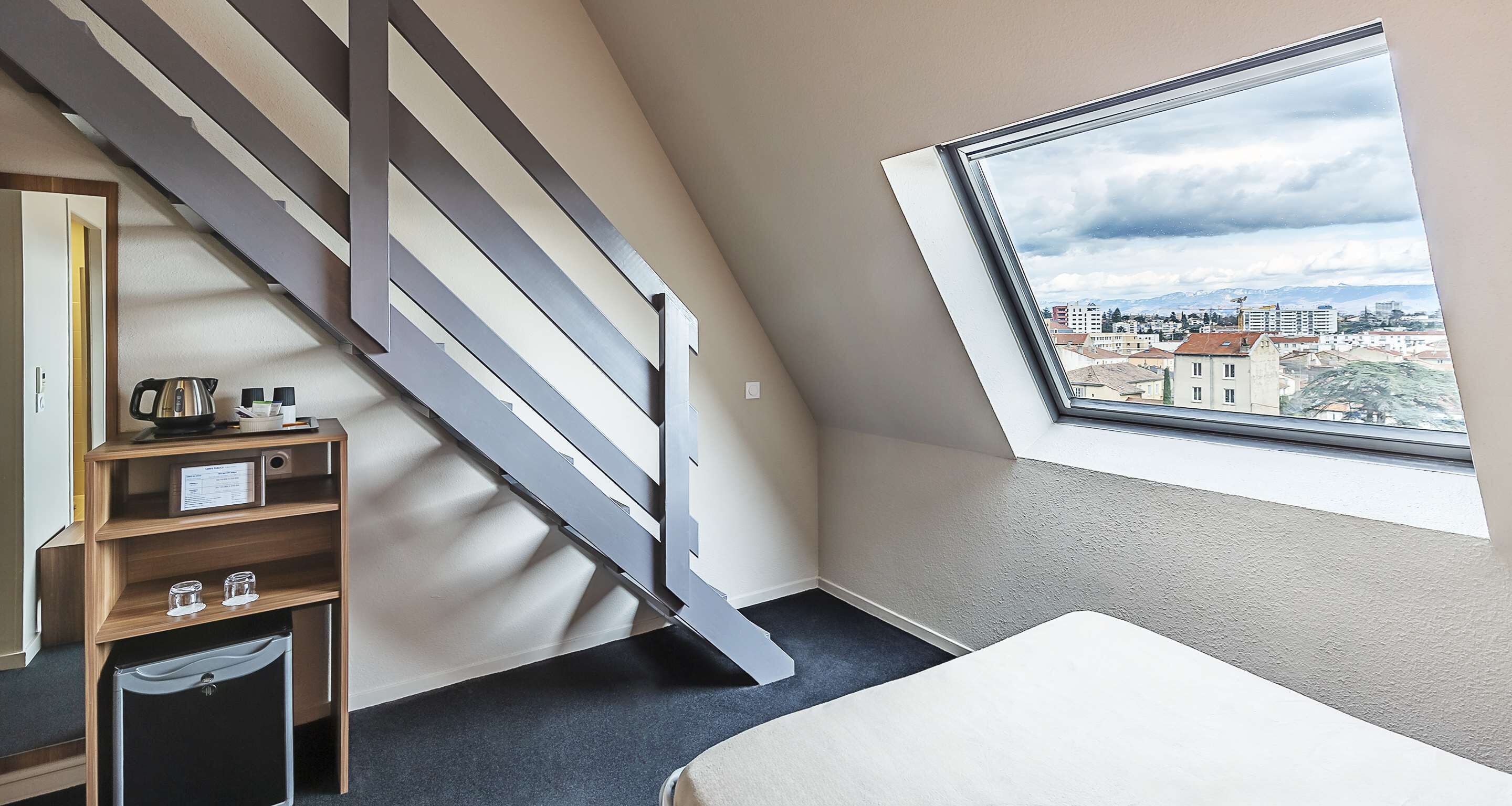 Een slaapkamer met een schuine kap, een ingebouwd bureau met waterkoker, een kleine koelkast en een raam met uitzicht op de stad.