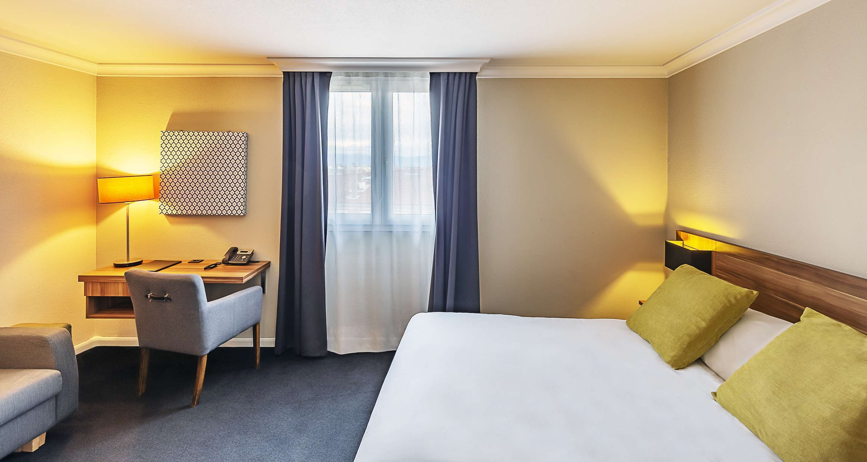 Et hotelværelse med seng, skrivebord, stol, lampe og udsigt over byen fra vinduet.