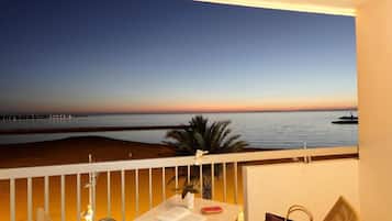A balcony with a view of the sea at sunset, a table with a book and a red object, and a chair.