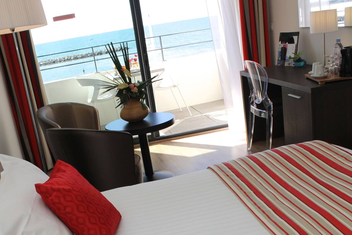 A hotel room with a bed, two red pillows, a desk with a lamp, and a view of the sea through large windows.