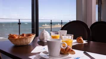 Ein Frühstückstisch mit Croissants, einem Teekännchen, Orangensaftgläsern und einer Tasse, mit Blick auf die Küste.