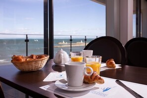 Una mesa de desayuno con cruasanes, un hervidor de agua, vasos de jugo de naranja y una taza, con vistas a la costa.