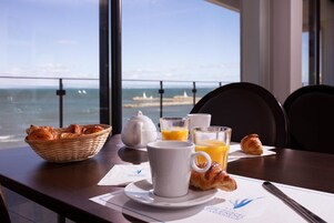 Una mesa de desayuno con cruasanes, un hervidor de agua, vasos de jugo de naranja y una taza, con vistas a la costa.