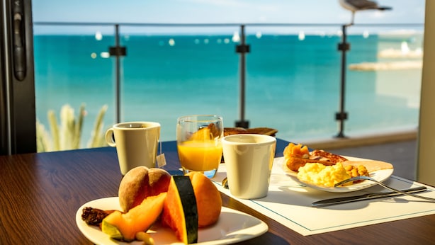 A breakfast table with a plate of food, two cups, and a glass of orange juice. A seagull is perched on a railing in the background. - Splendid Hôtel Camargue (Le Grau-Du-Roi)