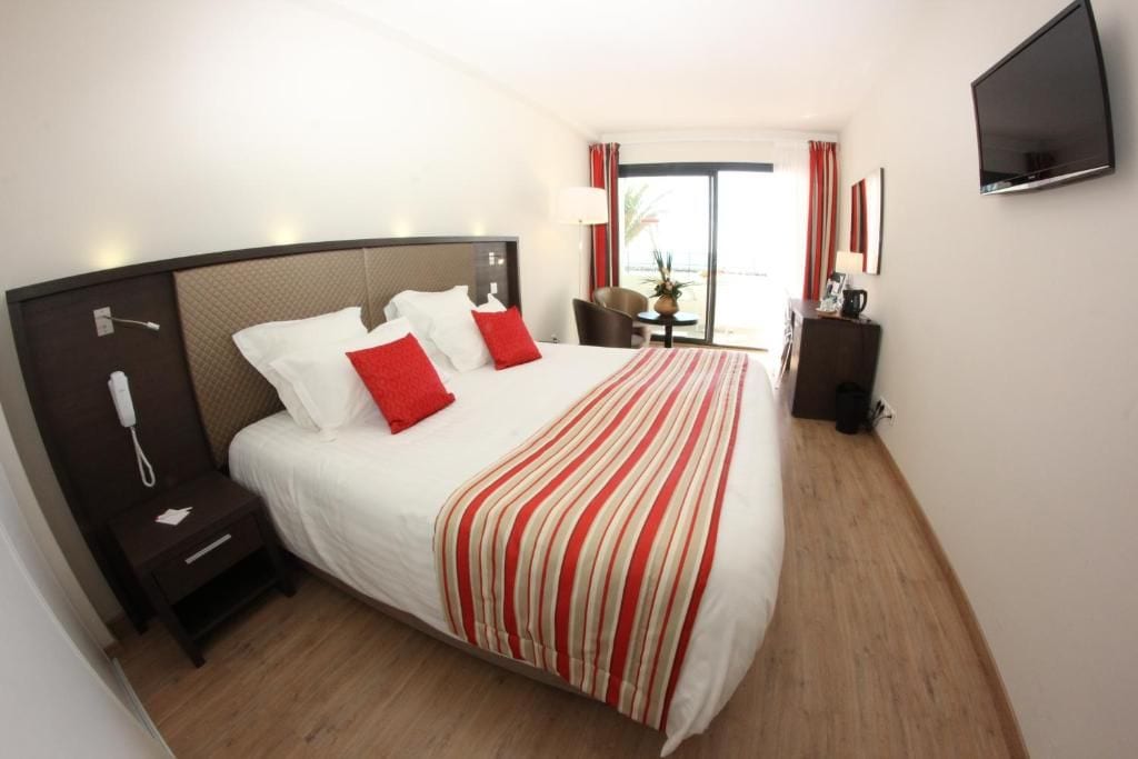 A hotel room with a bed, bedside table, lamp, TV, and a view of the outdoors.