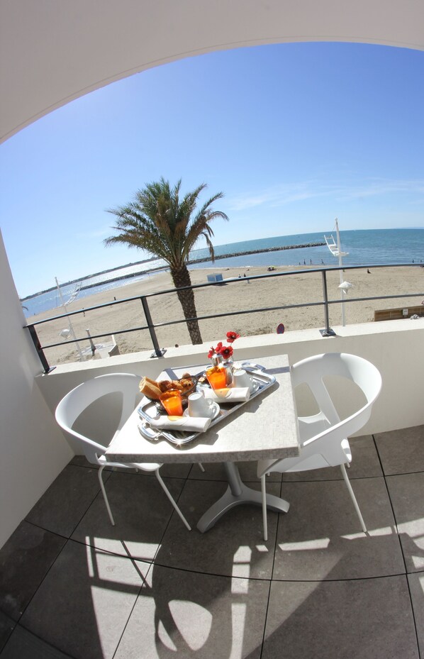 Un balcón con una mesa lista para el desayuno, con vista a una playa con una palmera y el mar.