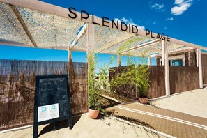Un espacio al aire libre techado con un letrero que dice 'Splendid Plage', plantas de bambú y una pizarra con información.