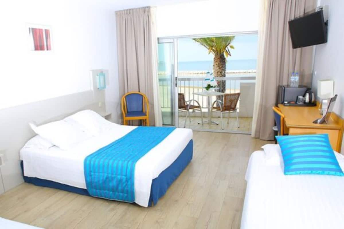 A hotel room with a bed, a desk, a chair, a TV, and a balcony with a view of the ocean.