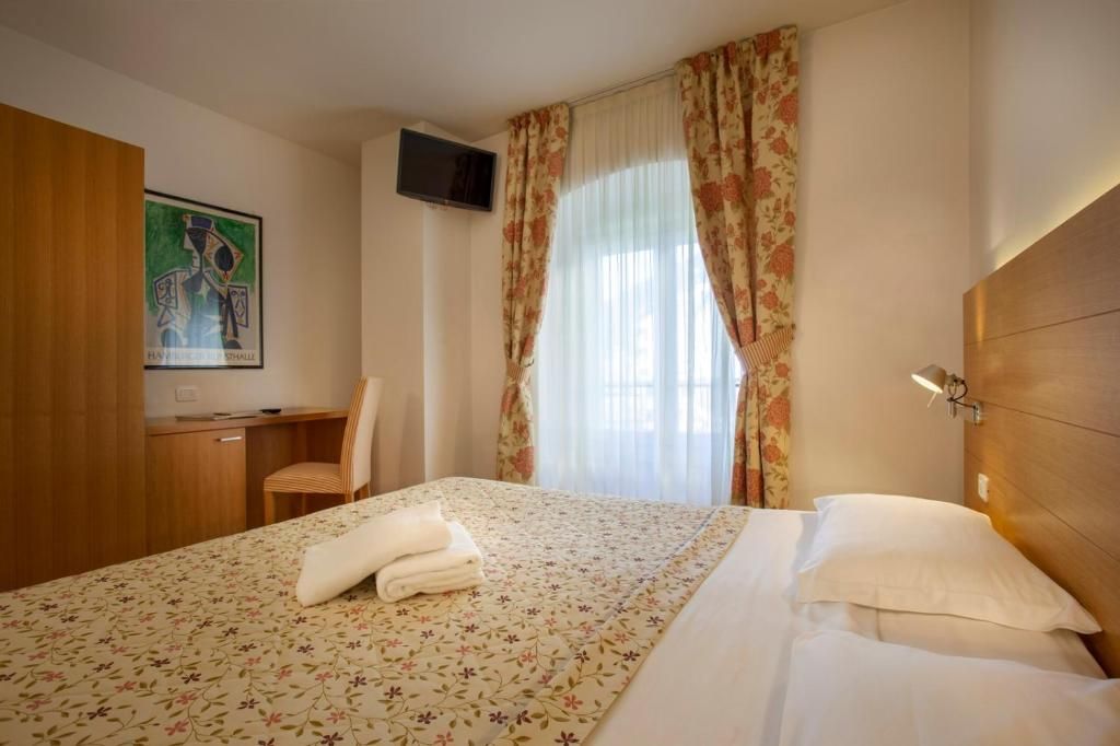 A hotel room with a bed, a desk, a chair, a television, and a painting on the wall.