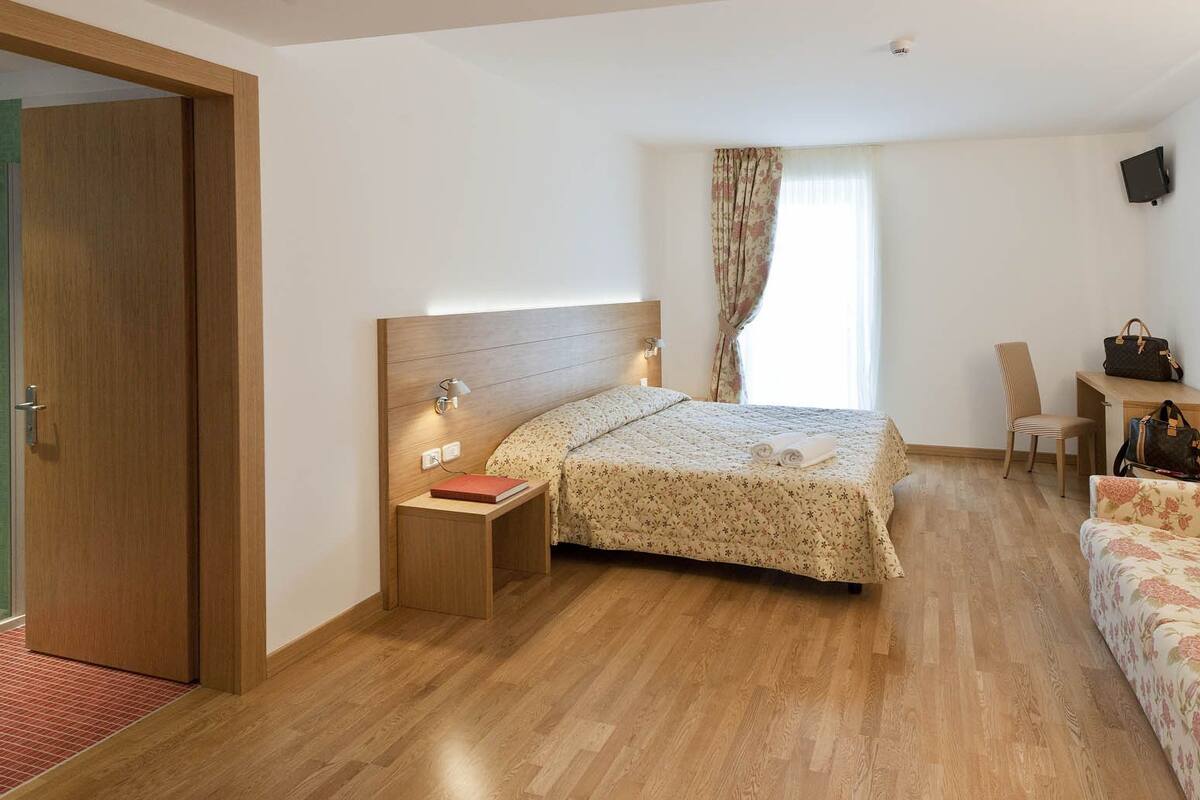 A hotel room with a wooden floor, a bed with a floral bedspread, a nightstand with a book, a chair, a desk with a handbag, and a TV on the wall.