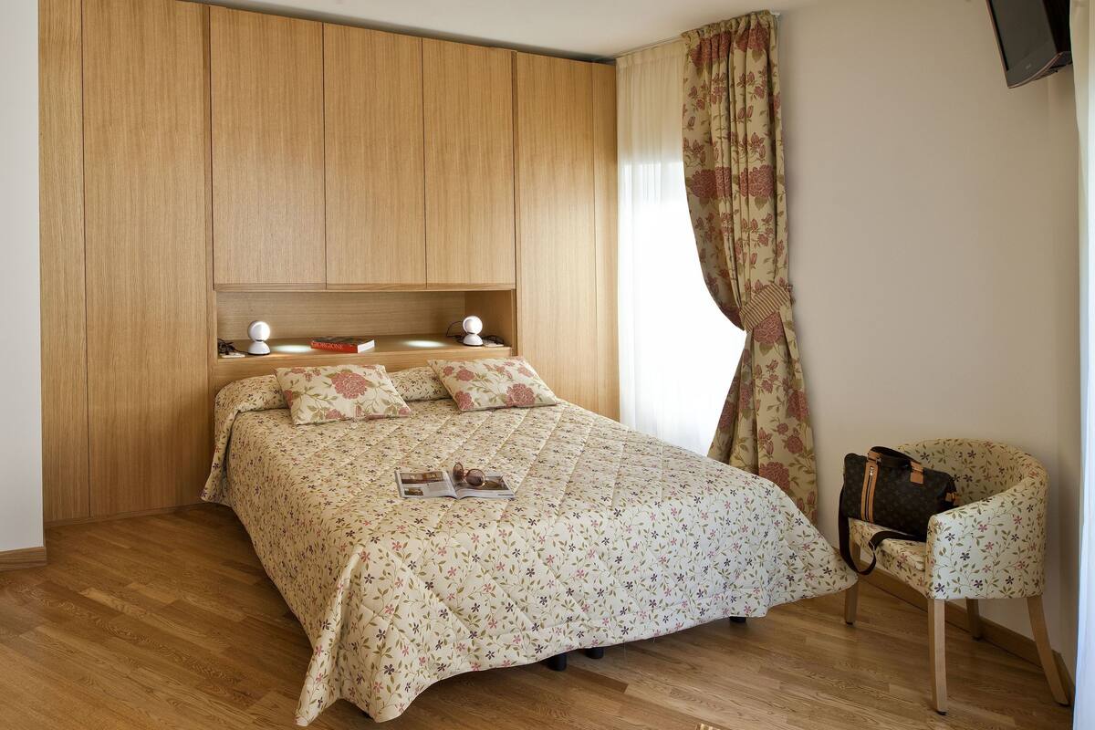 A bedroom with a wooden wardrobe, a bed with floral bedding, a chair, and a bathroom visible through an open door.