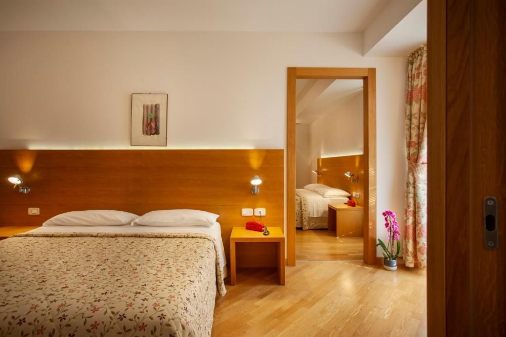 Habitación de hotel con cabecera de madera, cama con ropa blanca, mesita de noche con lámpara y un espejo que refleja la habitación.