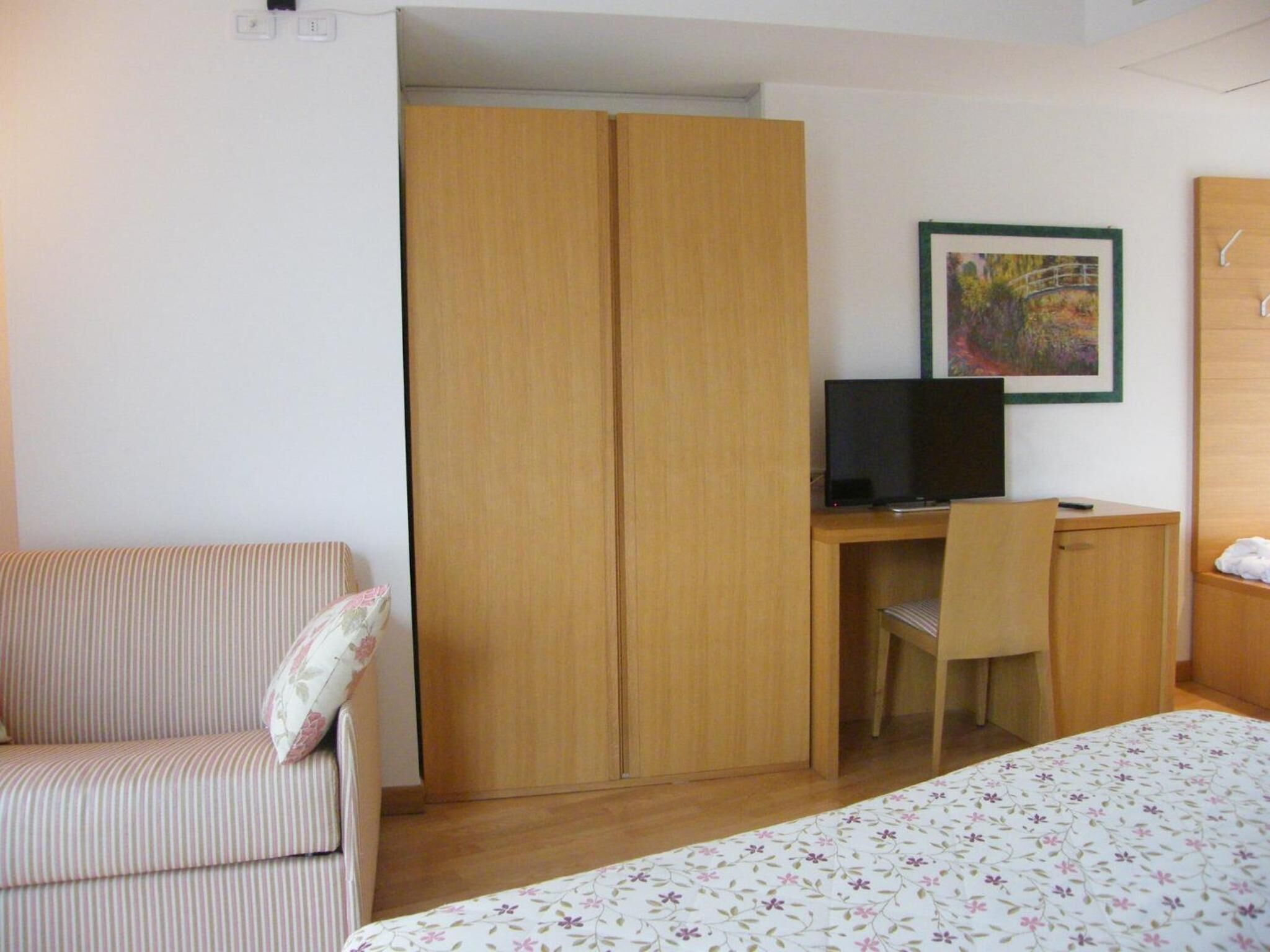 A hotel room with a striped sofa, a wooden wardrobe, a desk with a television, and a bed with a floral bedspread.