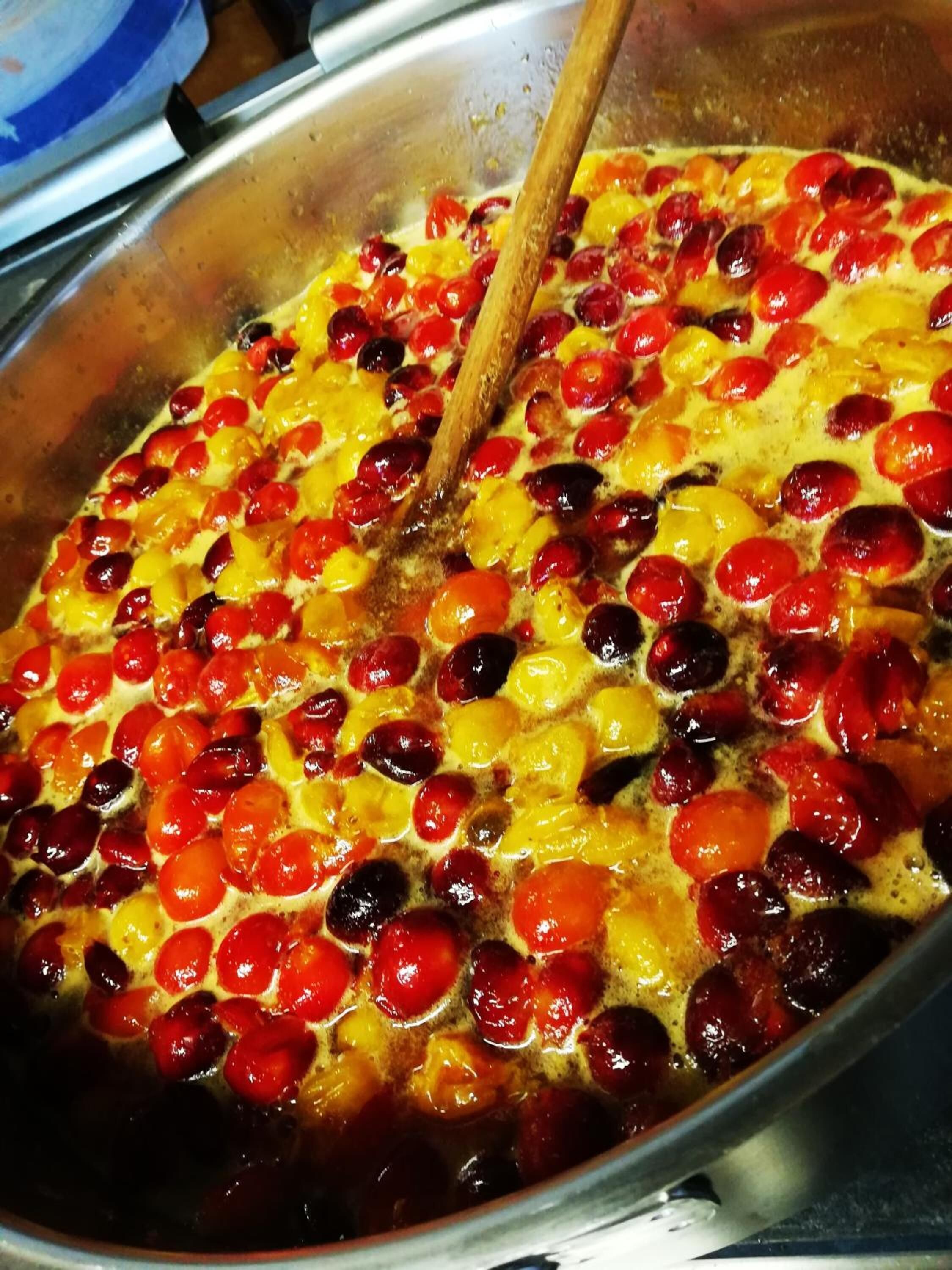 A large pot with a wooden spoon, containing a mixture of red and yellow fruits, possibly cherries and apricots, being cooked or mixed.