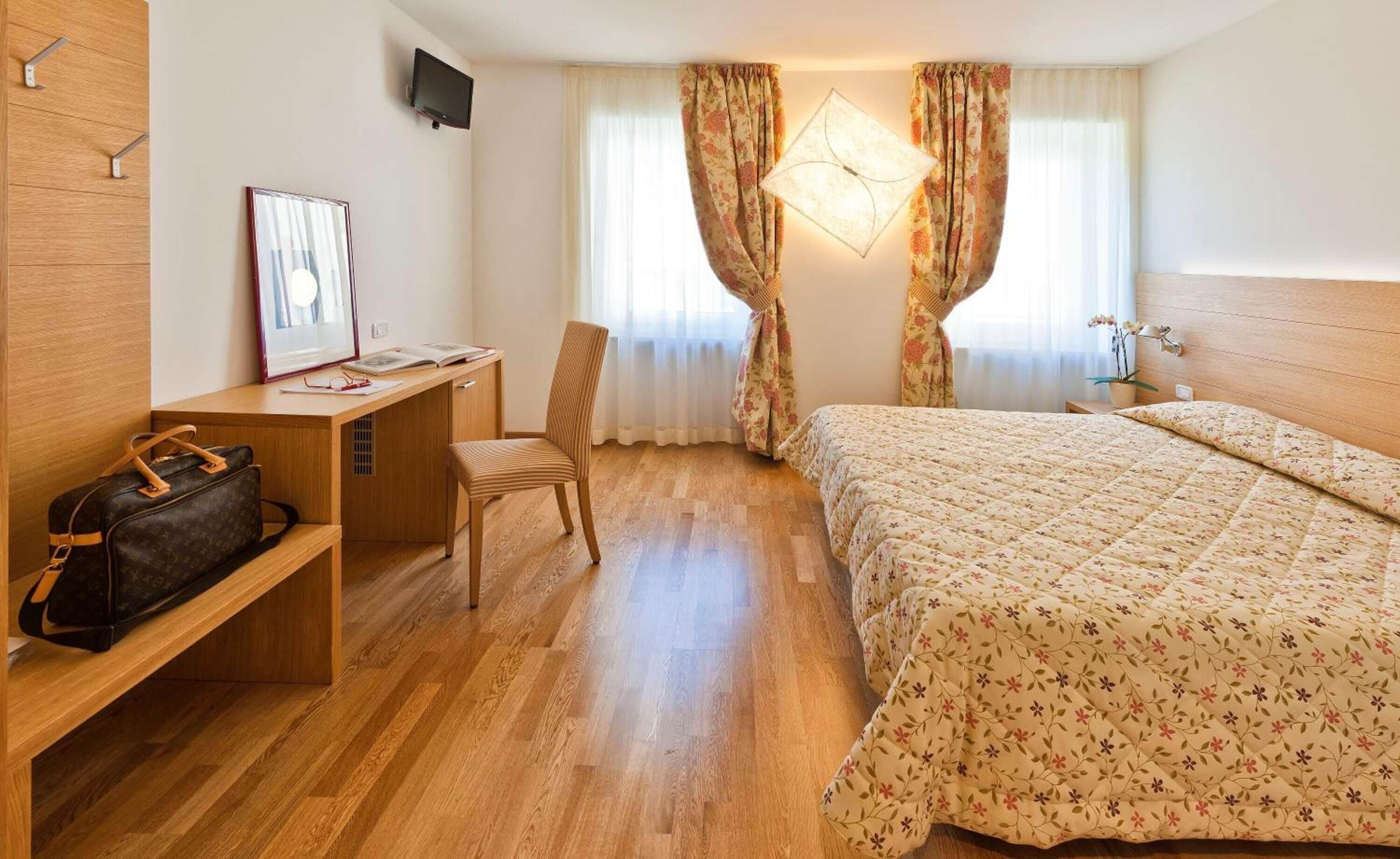 Una habitación de hotel con escritorio de madera, una silla, una cama con ropa de cama floral y una mesita de noche con lámpara.
