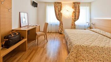 Una habitación de hotel con escritorio de madera, una silla, una cama con ropa de cama floral y una mesita de noche con lámpara.