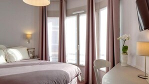 A hotel room with a large bed, a desk, a chair, a TV mounted on the wall, and a window with curtains. - Ours Blanc-Wilson (Toulouse)