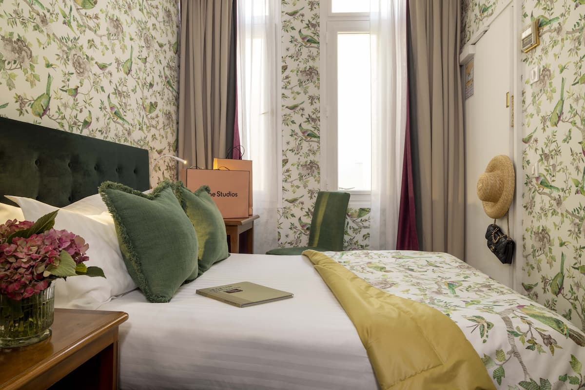 Un lit bien fait, recouvert d’une couvre-lit à motifs floraux, d’un plaid vert et agrémenté d’un vase rempli de fleurs roses, posé sur une table de chevet en bois.
