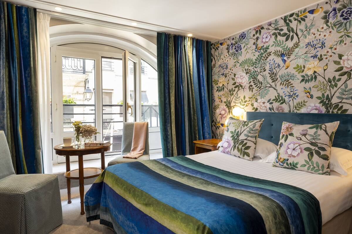 Une chambre d’hôtel avec un lit, une chaise, une petite table et un balcon avec des rideaux.