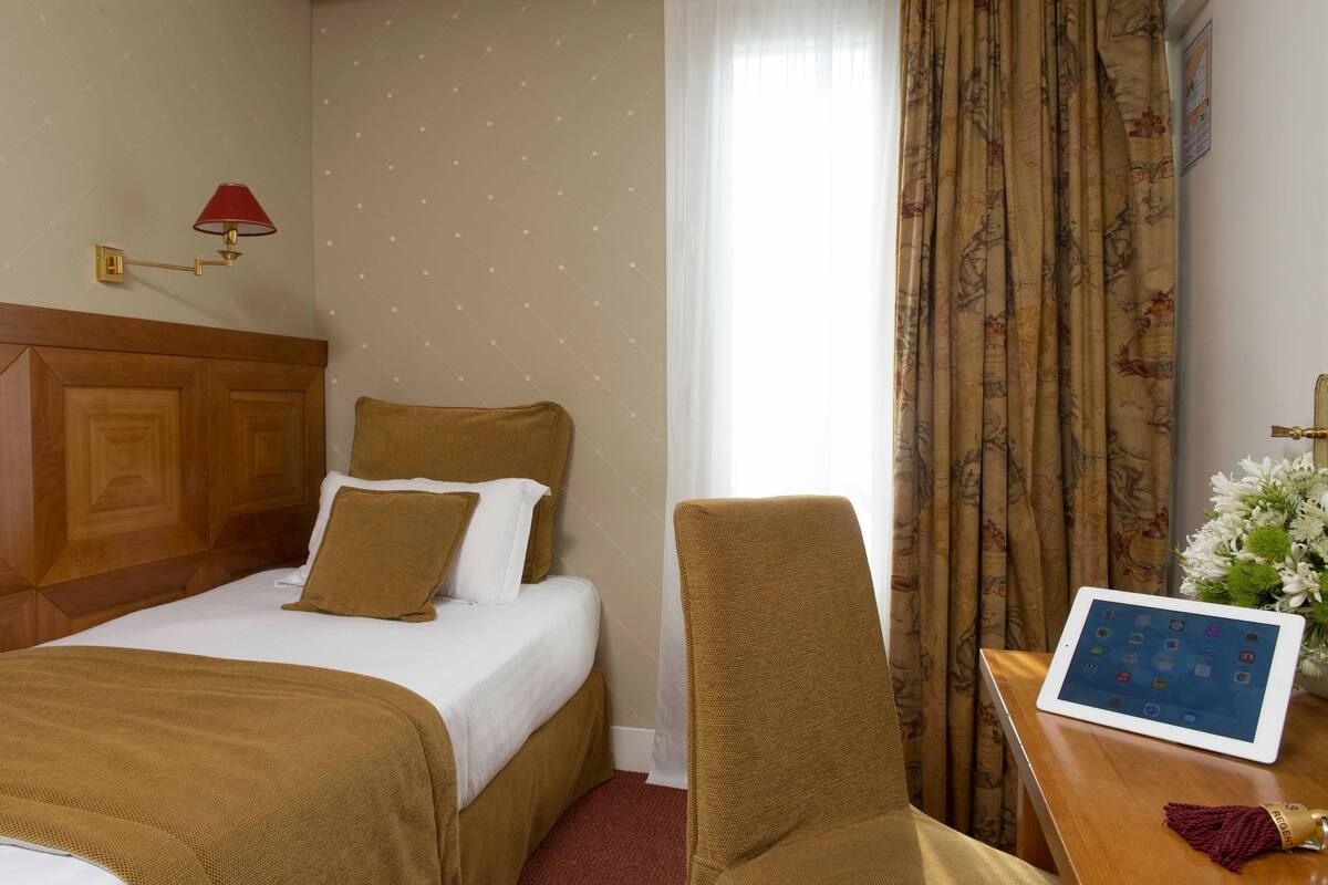 Une chambre d’hôtel avec un lit, une chaise, une table sur laquelle se trouve une tablette, une lampe et un rideau.
