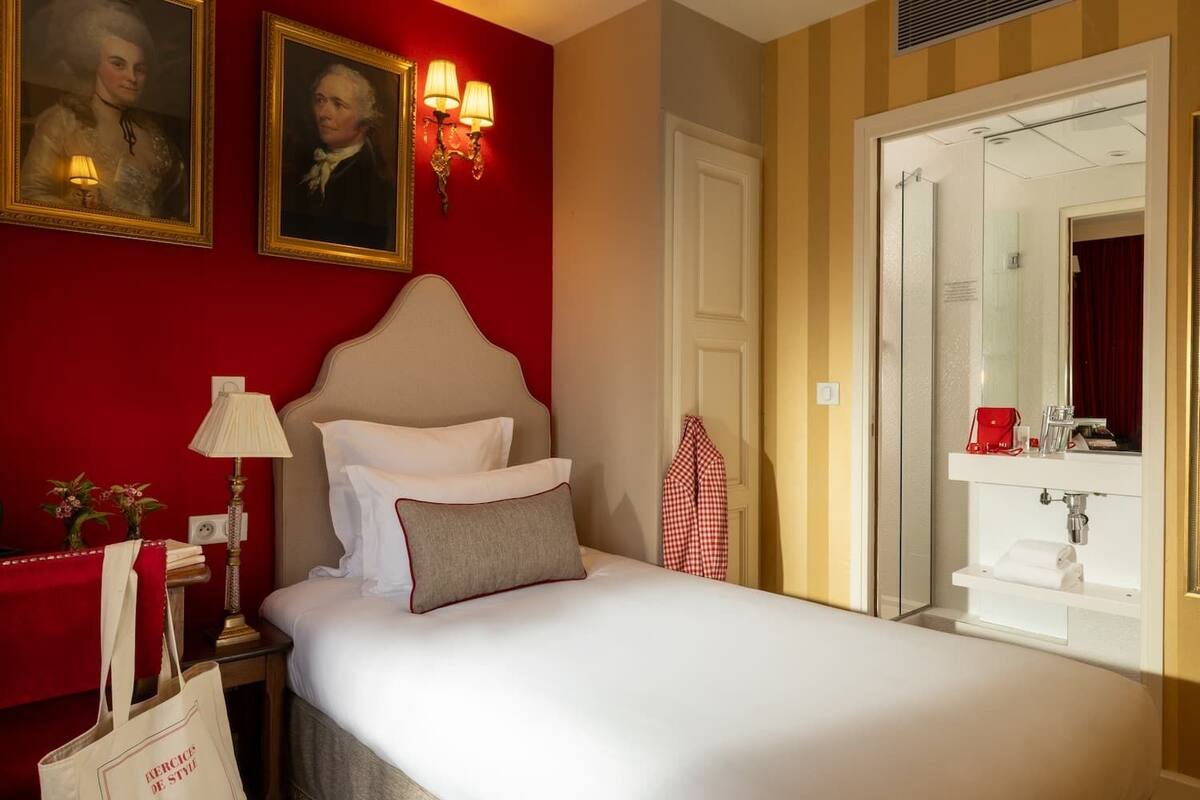 A hotel room with a bed, a nightstand with a lamp, a red wall with two framed portraits, a red and white checkered towel, and a bathroom with a mirror and sink.