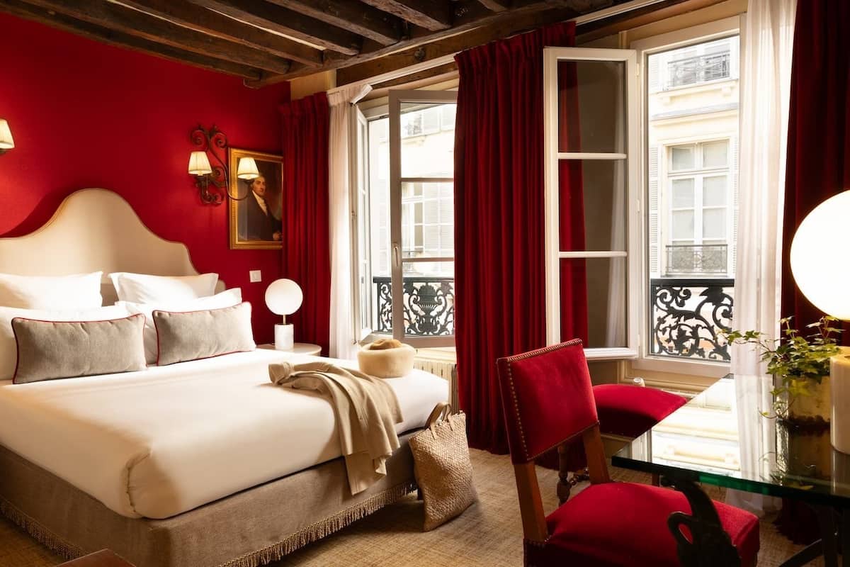 A hotel room with a bed, a chair, a table with a lamp, a window with curtains, and a view of the outside.