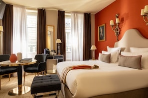 Quarto de hotel com uma cama grande, uma escrivaninha, uma cadeira, uma mesa redonda e uma janela com cortinas.