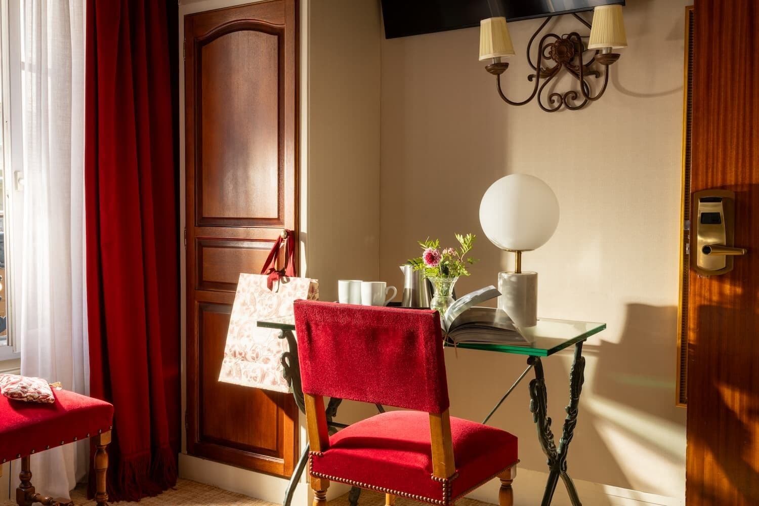 A room with a wooden door, a red chair, a glass table with a book, a vase with flowers, and a lamp.