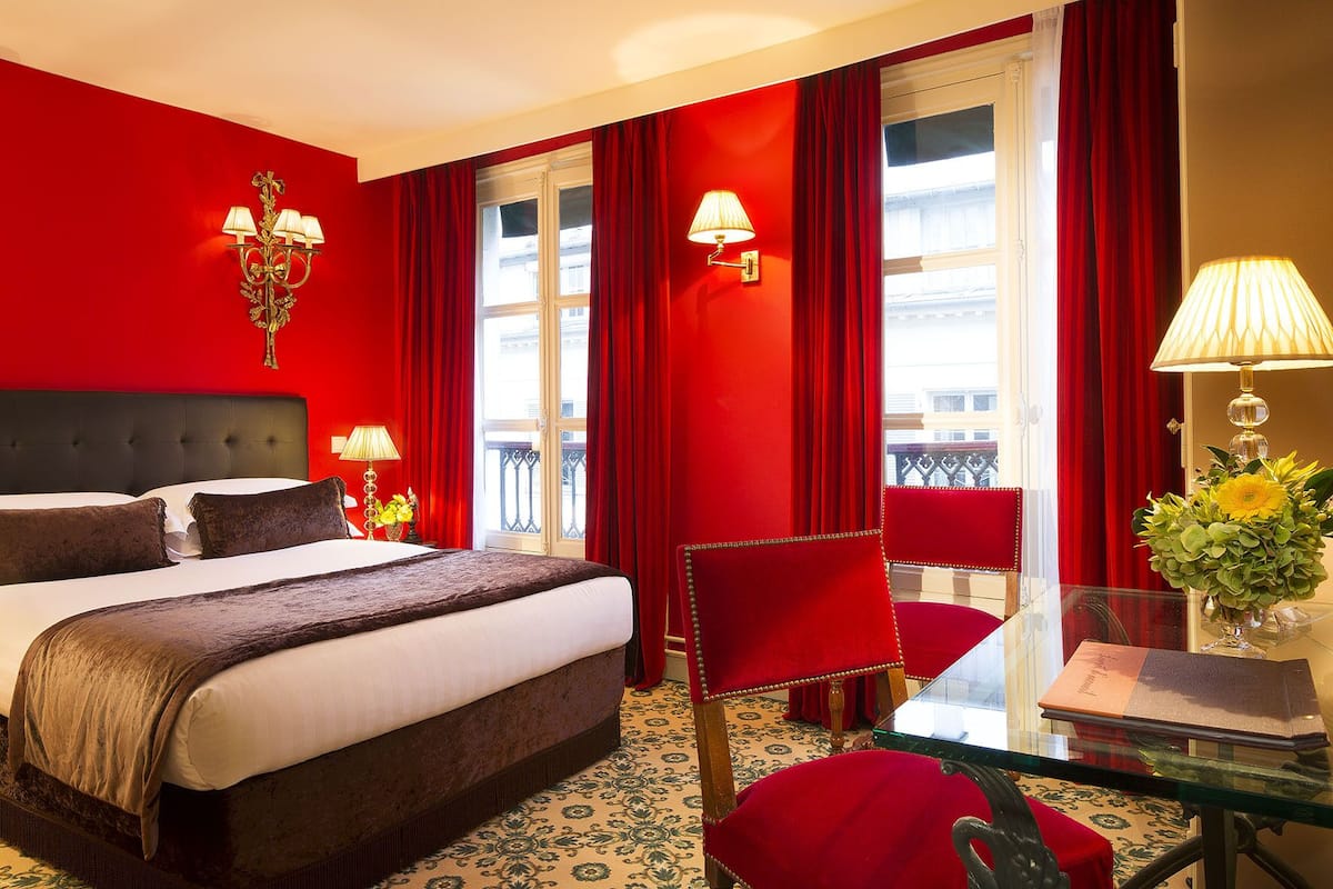 A hotel room with a large bed, a desk with a chair, red curtains, and a window with a view.