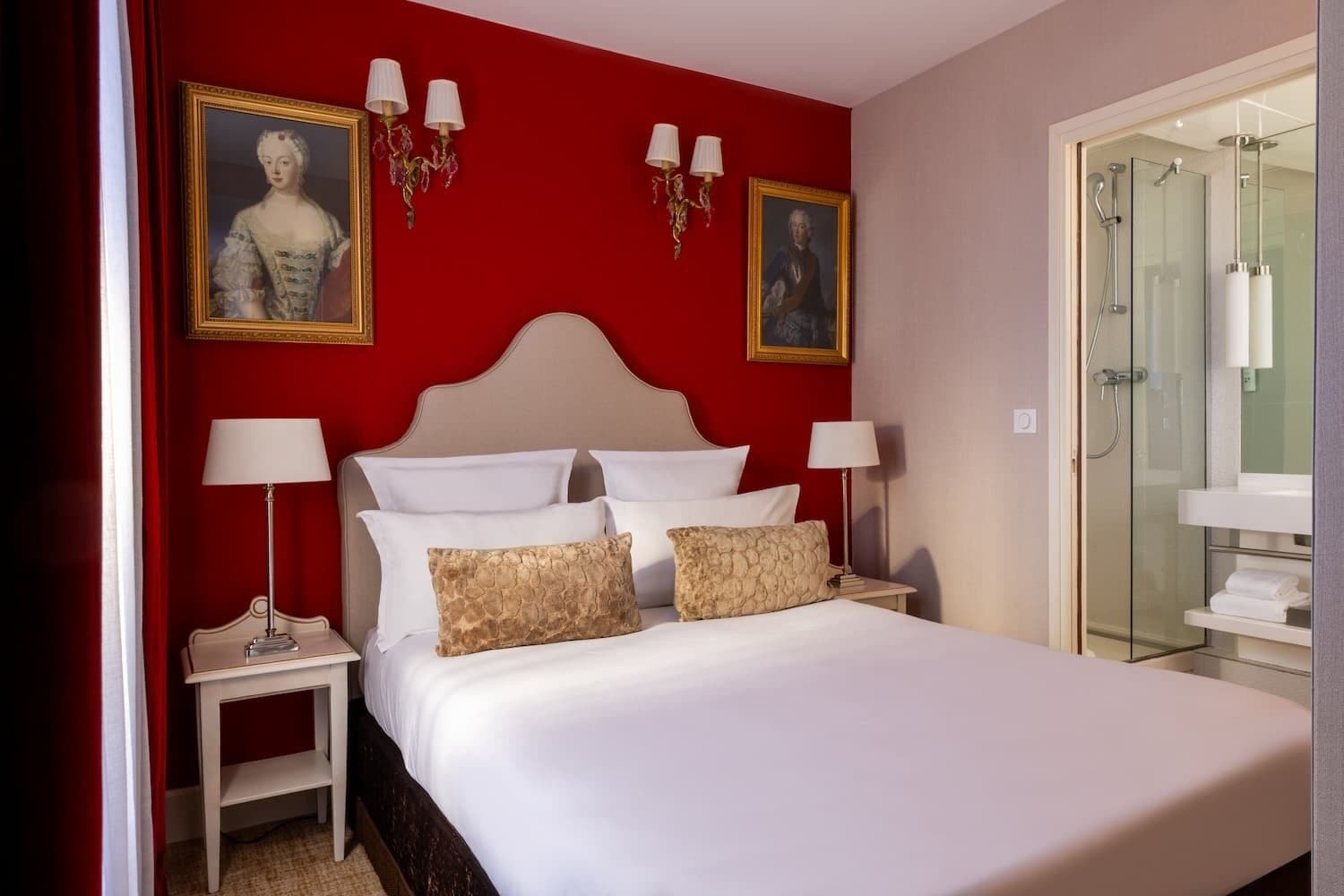 Une chambre d’hôtel avec un mur d’accent rouge, un grand lit avec du linge de lit blanc, deux tables de chevet avec des lampes et une cabine de douche en verre.